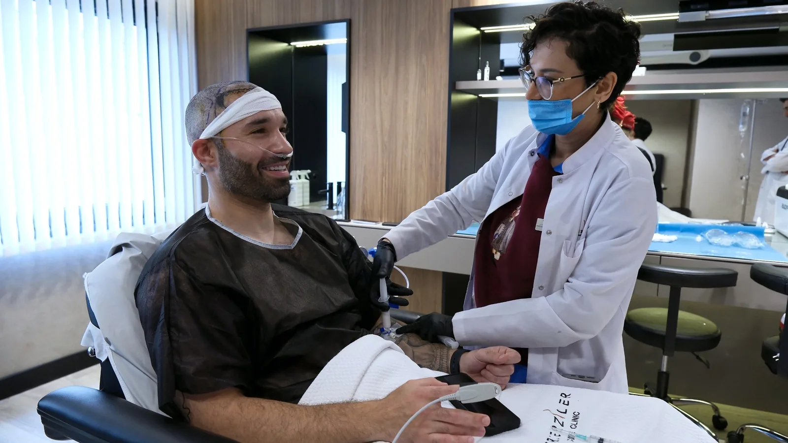 Smiling hair transplant patient during IV sedation while Dr. Semra Uludemir checks his IV line, with a pulse monitor on his finger, at Dr. Terziler Exclusive Clinic, Istanbul.
