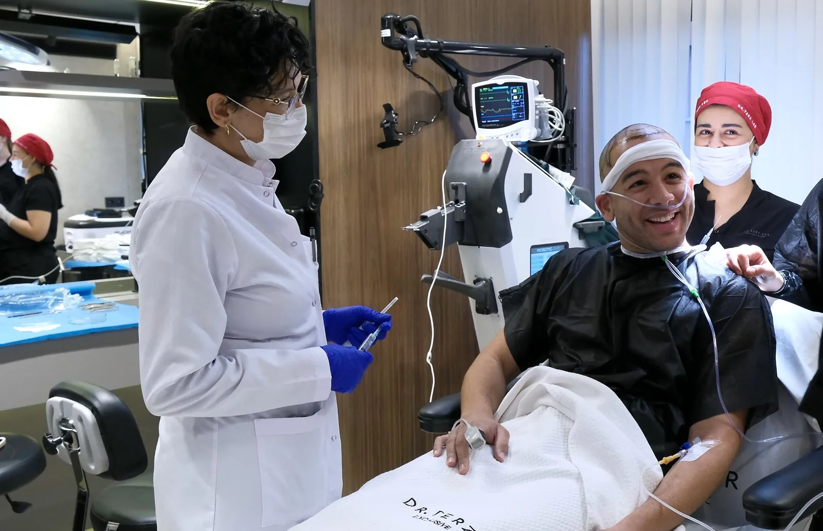 Hair transplant patient smiling under IV sedation in the treatment chair, hairline marked and vital signs monitored, beside the Robotic DHI machine at Dr. Terziler Exclusive Clinic, Istanbul.